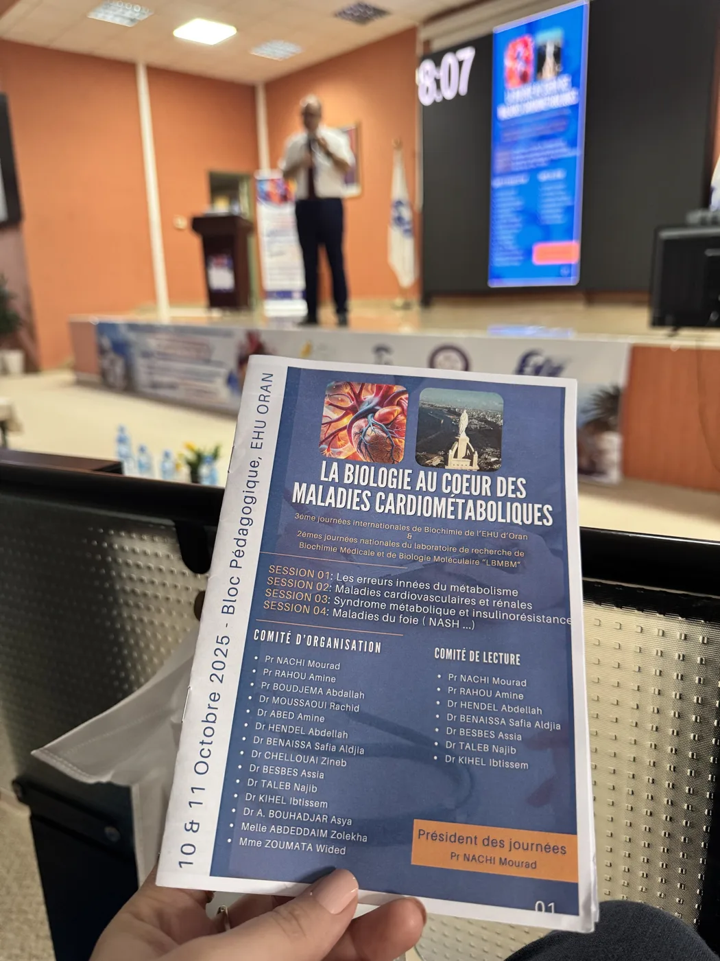 Programme imprimé du congrès tenu en main, devant la scène et l'écran du congrès
